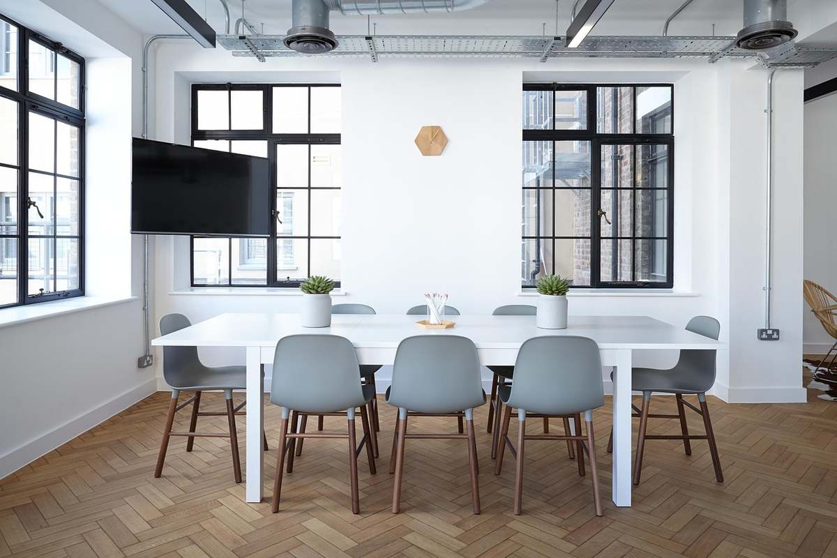 Salle de réunion avec table blanche, six chaises gris, deux plantes, deux fenêtres, sol en parquet